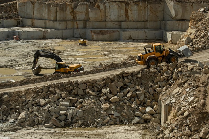 A quarry site showcasing heavy machinery extracting stone from the earth, surrounded by rocky terrain and dust clouds.