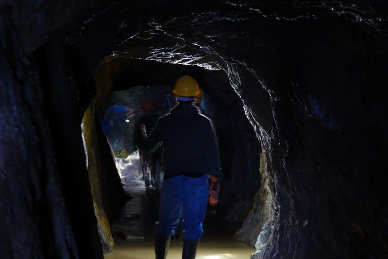 A miner operates machinery in a dimly lit underground tunnel, surrounded by rock formations and mining equipment.